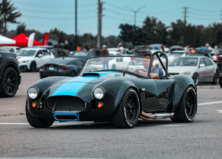 A classic black and blue Cobra car driven by a man at an outdoor event with other cars in the background.