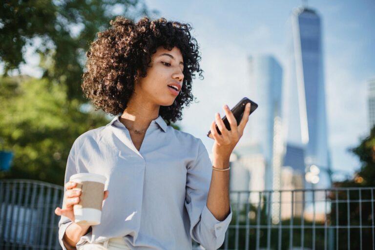 Young woman with curly hair uses smartphone outdoors, holding coffee.