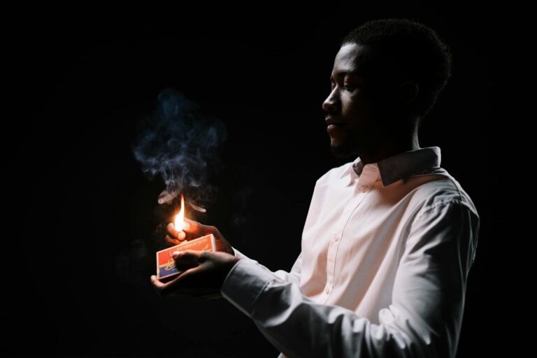 African American man in white shirt igniting a matchstick, casting warm light in darkness.
