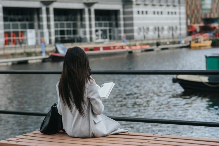 Back view of unrecognizable female in warm clothes reading book at spare time on waterfront