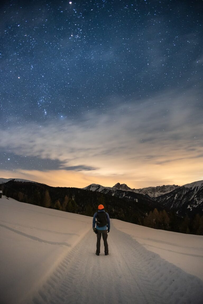 A lone hiker gazes at the starry sky atop a snowy mountain trail, evoking a sense of adventure and tranquility.