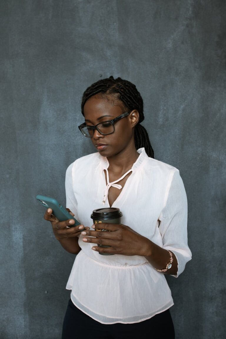 African American woman in glasses holds a coffee and smartphone, focused indoors.