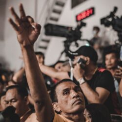A man raises his hand in a crowd during an event in Bangkok, Thailand, with cameras in the background.