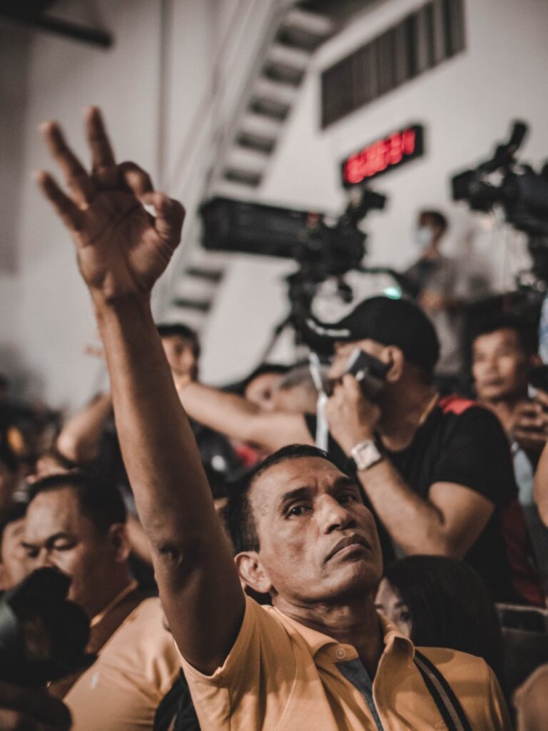 A man raises his hand in a crowd during an event in Bangkok, Thailand, with cameras in the background.
