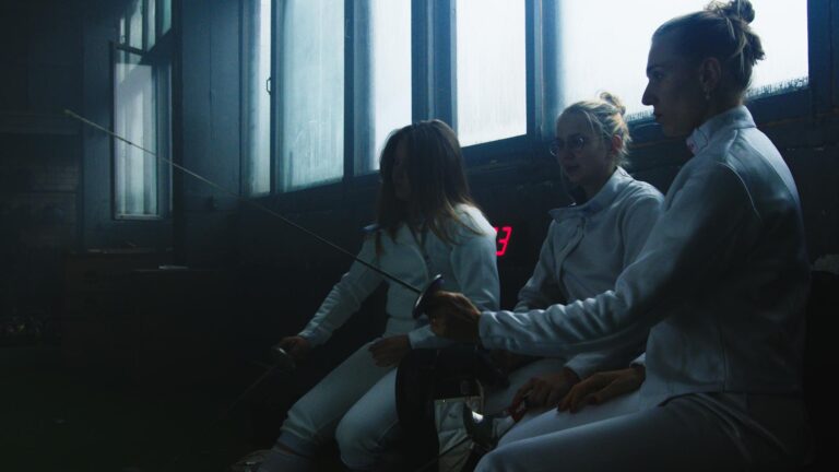 Three women fencers in white uniforms sit indoors, ready for training.