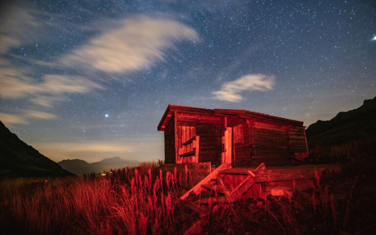 A wooden cabin illuminated by red light under a starry night sky in a rural landscape.