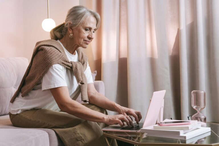 An elderly woman sits comfortably indoors using a laptop, highlighting modern technology use among seniors.