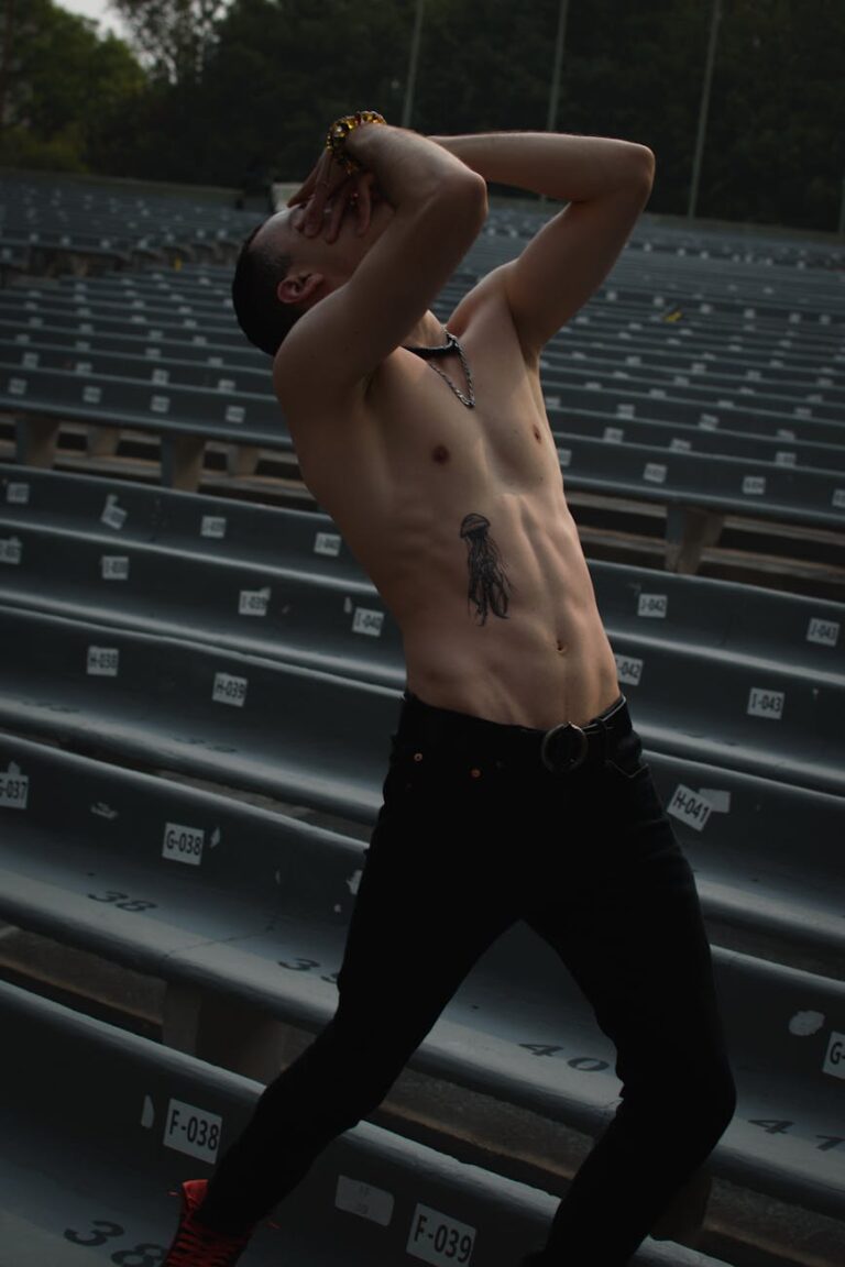 Athletic man posing shirtless, showing tattoos, in a deserted outdoor auditorium setting.