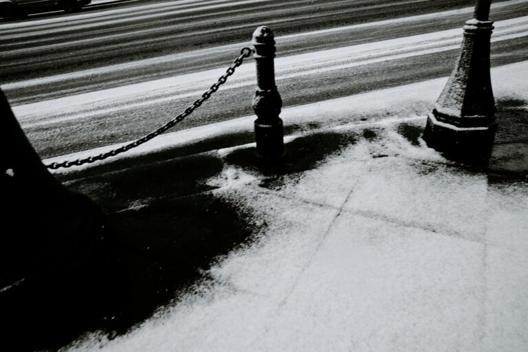 Black and white image of a snowy, urban street with chain posts. Quiet winter scene.