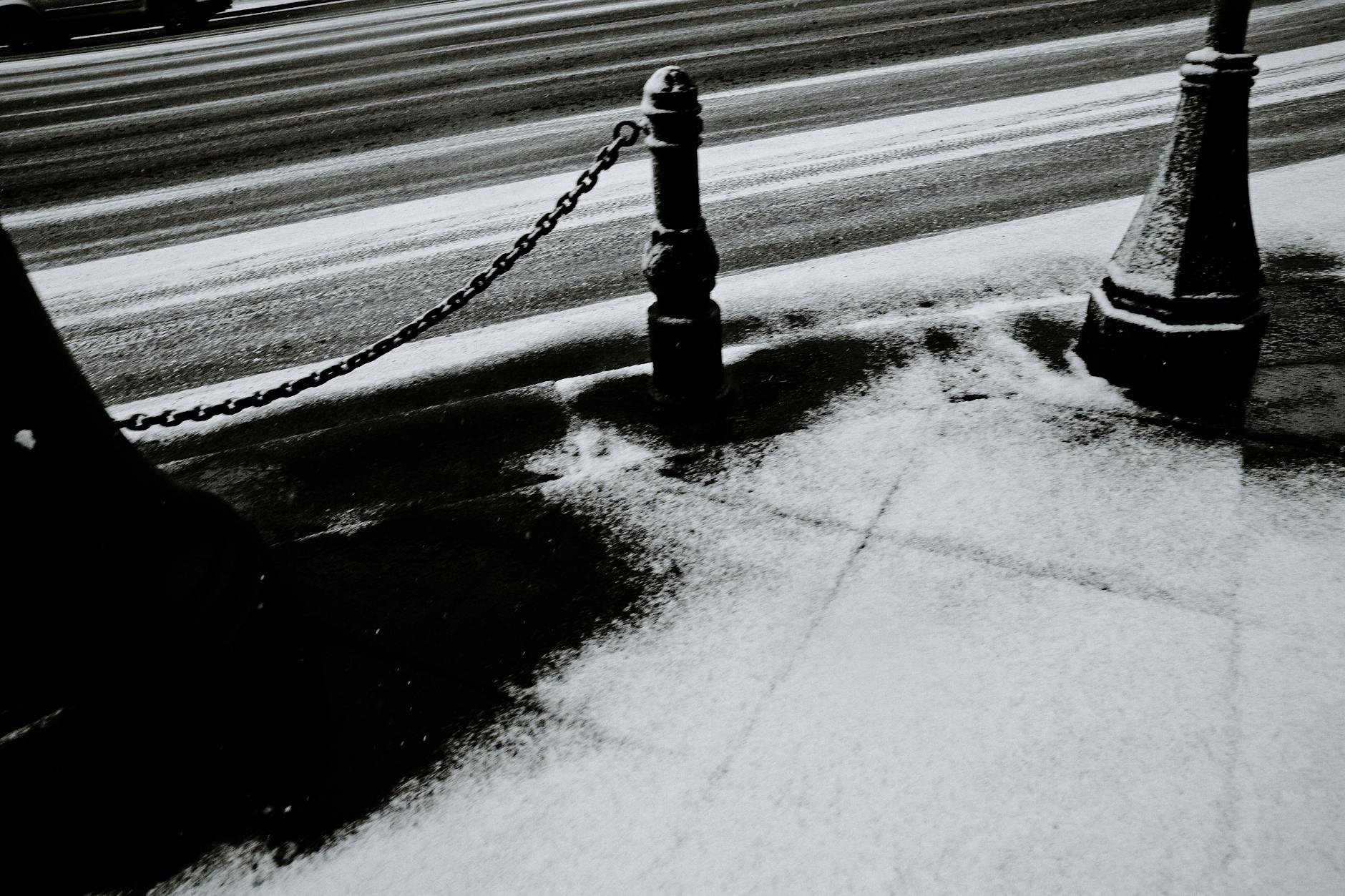 Black and white image of a snowy, urban street with chain posts. Quiet winter scene.