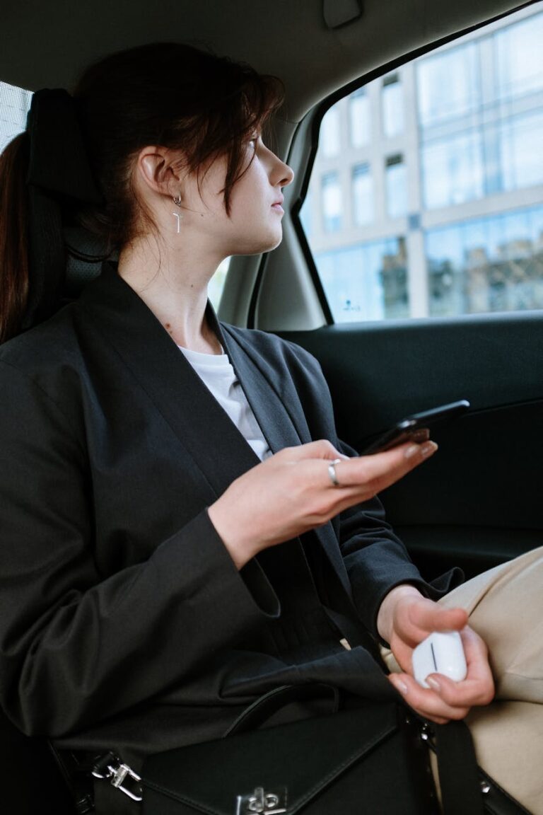 A woman with a ponytail and earrings uses a smartphone in the back of a car, focusing outside.