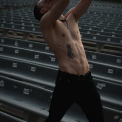 Athletic man posing shirtless, showing tattoos, in a deserted outdoor auditorium setting.
