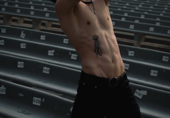 Athletic man posing shirtless, showing tattoos, in a deserted outdoor auditorium setting.
