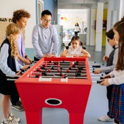 A diverse group of children and a young adult playing foosball together indoors.