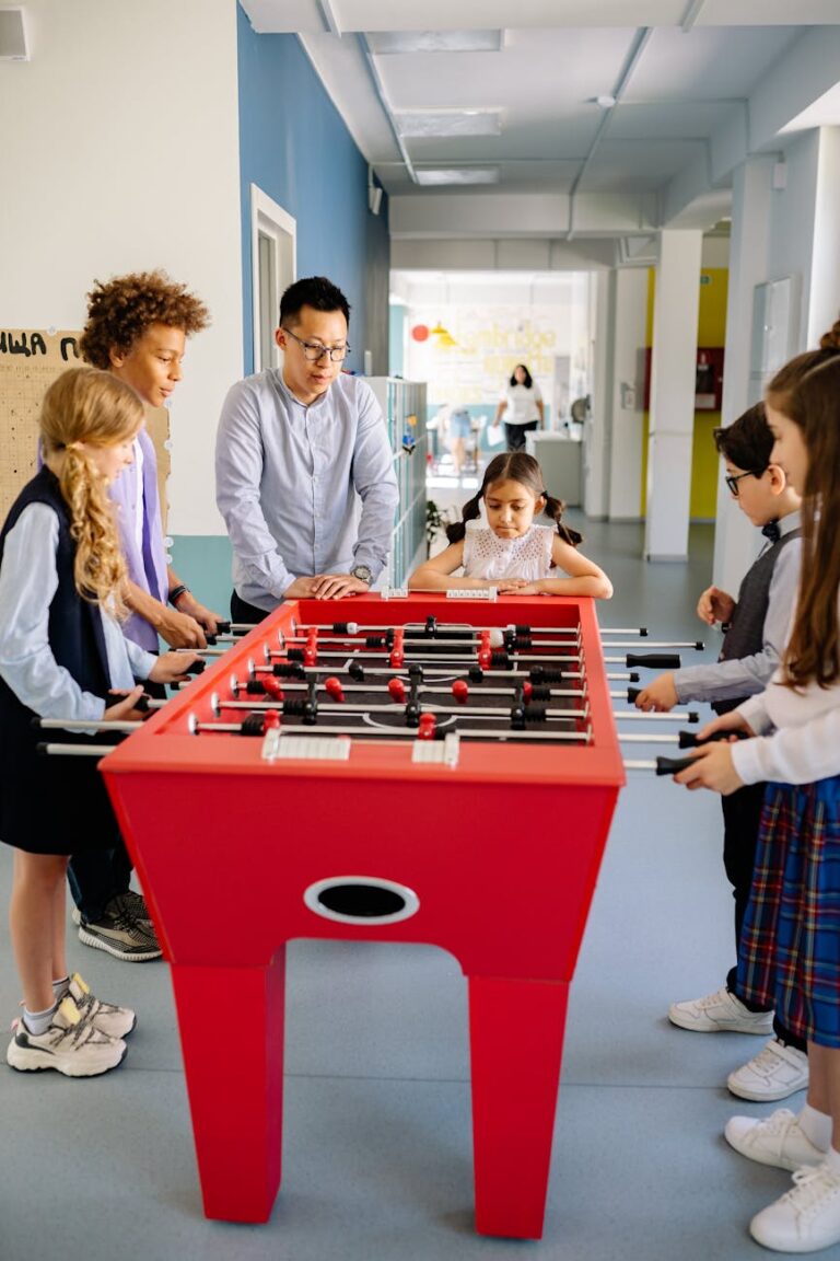 A diverse group of children and a young adult playing foosball together indoors.