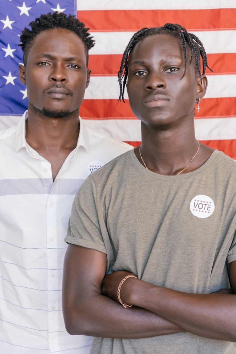Two black men with Vote stickers stand before an American flag, symbolizing civic engagement.