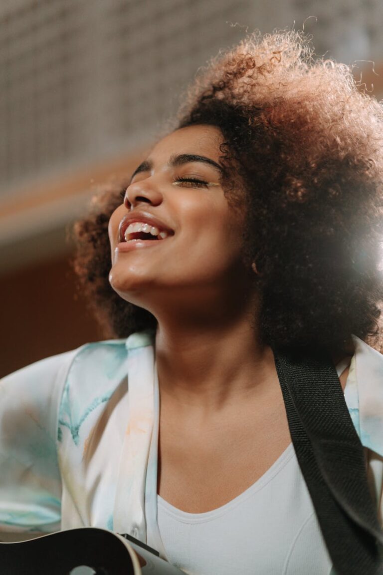 Smiling woman with afro hair sings joyfully while playing guitar indoors.