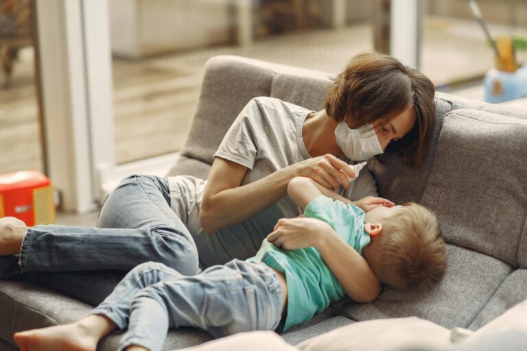 A mother wearing a face mask comforts her sick son with nasal drops on the sofa.