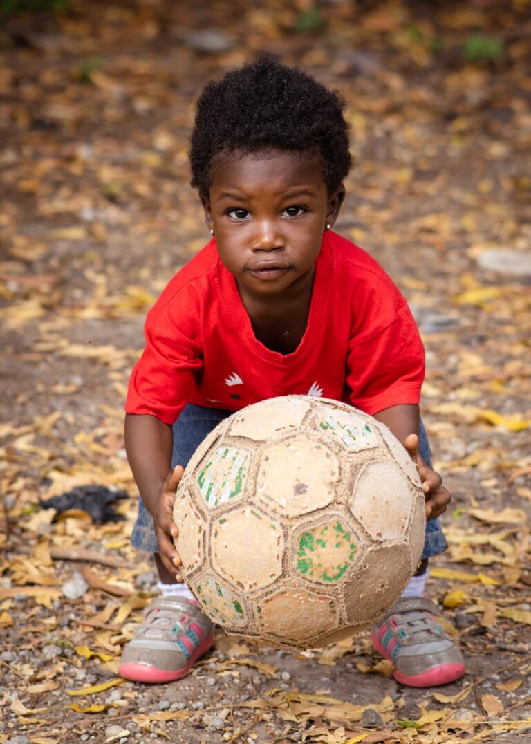 A young child in a red shirt holds a soccer ball outdoors, surrounded by leaves.
