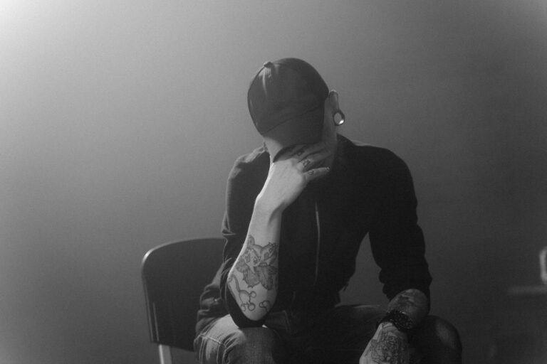 A tattooed person in a cap sits thoughtfully in a smoky room, conveying emotions and introspection.