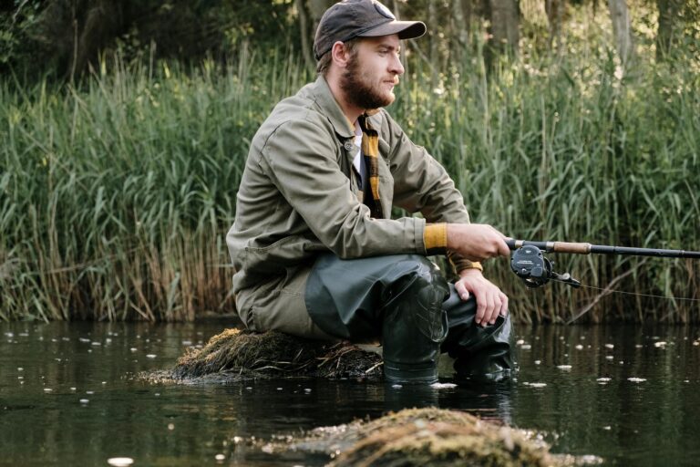 Bearded man in cap fishing by a river surrounded by lush reeds and nature.