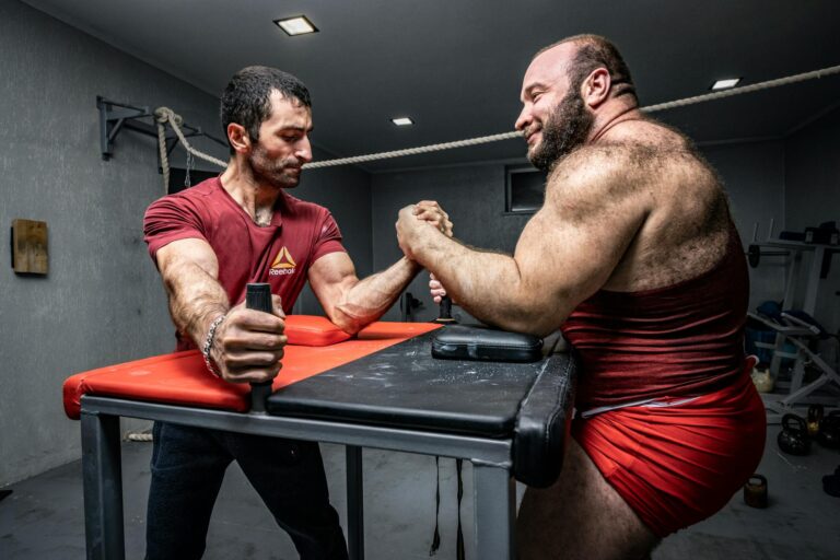 Two muscular men engaged in a competitive arm wrestling match indoors.