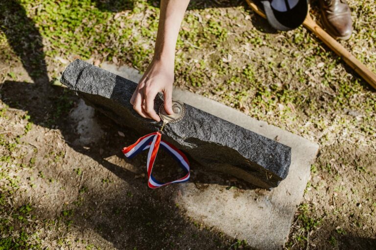 A hand placing a medal on a tombstone in a grassy cemetery, symbolizing honor and remembrance.