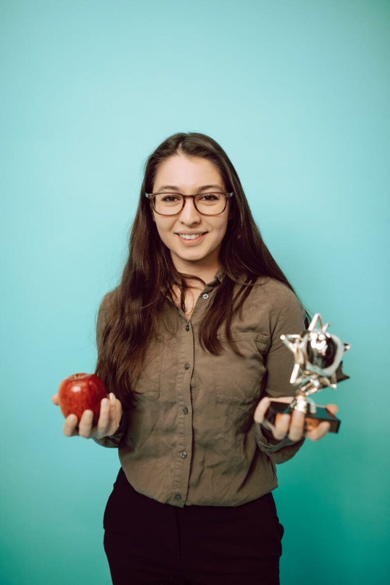 Smiling young woman in glasses holds an apple and a trophy on a blue background.