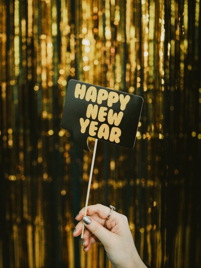 A festive hand holding a 'Happy New Year' sign against a golden curtain backdrop.