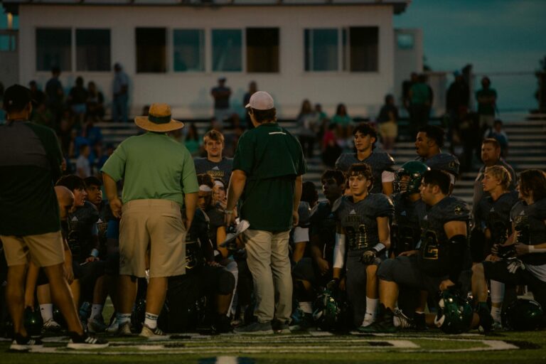 Football team huddles with coaches during a game at a stadium under the evening sky.
