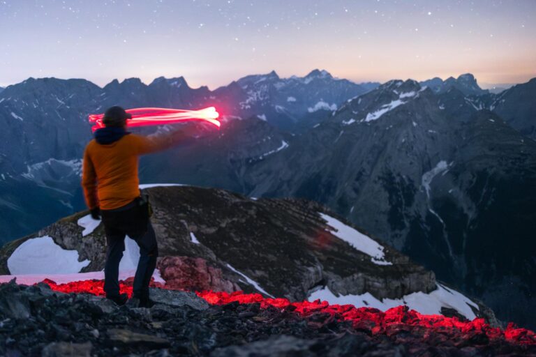 A lone hiker signals with red light atop a mountain at dusk, surrounded by snowy peaks.