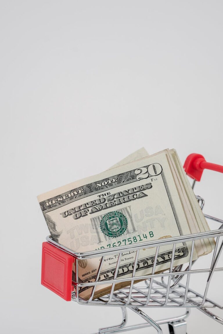 A stack of US dollar bills placed in a miniature shopping cart over a white background.