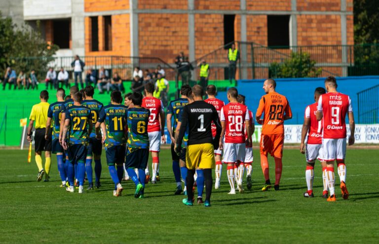 Players from two teams in blue and red uniforms preparing for a football match on a green field.