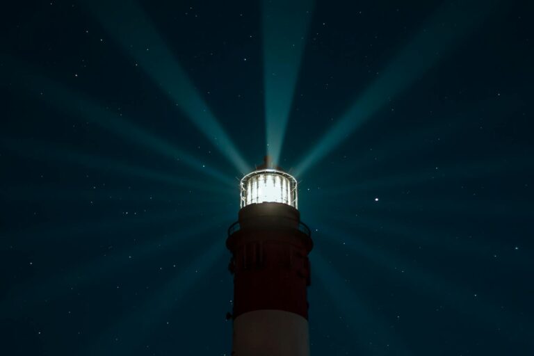 A lighthouse beams light across a starry night sky, creating an ethereal glow over the sea.
