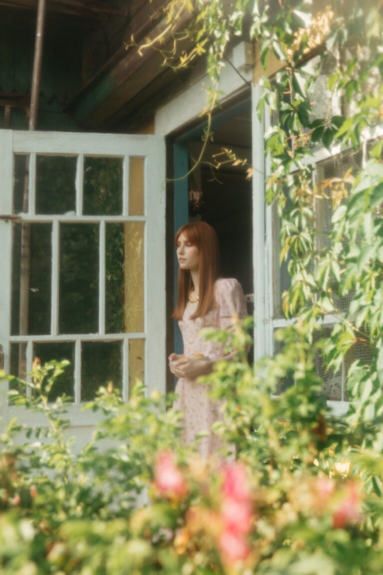 A woman in a printed dress stands by an open doorway surrounded by lush plants.