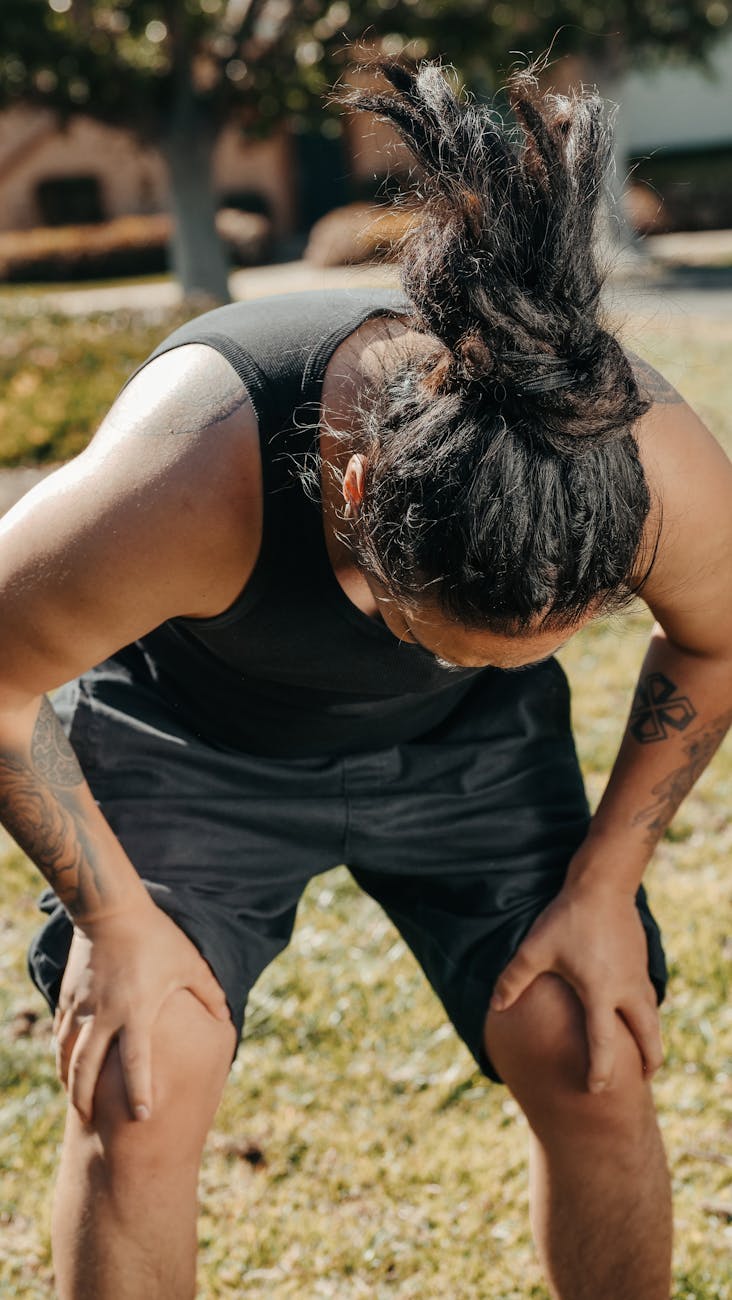 A man in a tank top holds his knees in apparent pain in a sunny park setting.